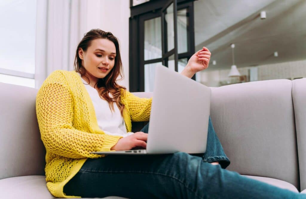 A woman in a yellow sweater sitting on the couch with her laptop working on her blog. Keep reading to learn how to monetize your blog from day one.