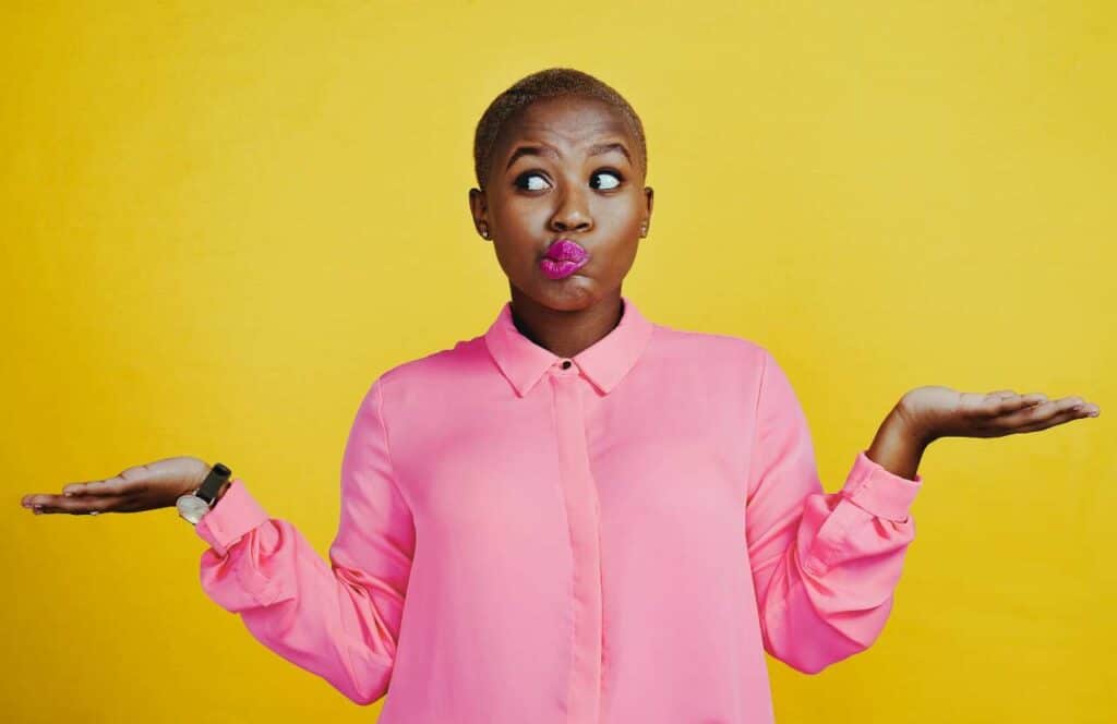 A black woman with pink lipstick and pink shirt standing with her palms facing up as if she is confused.