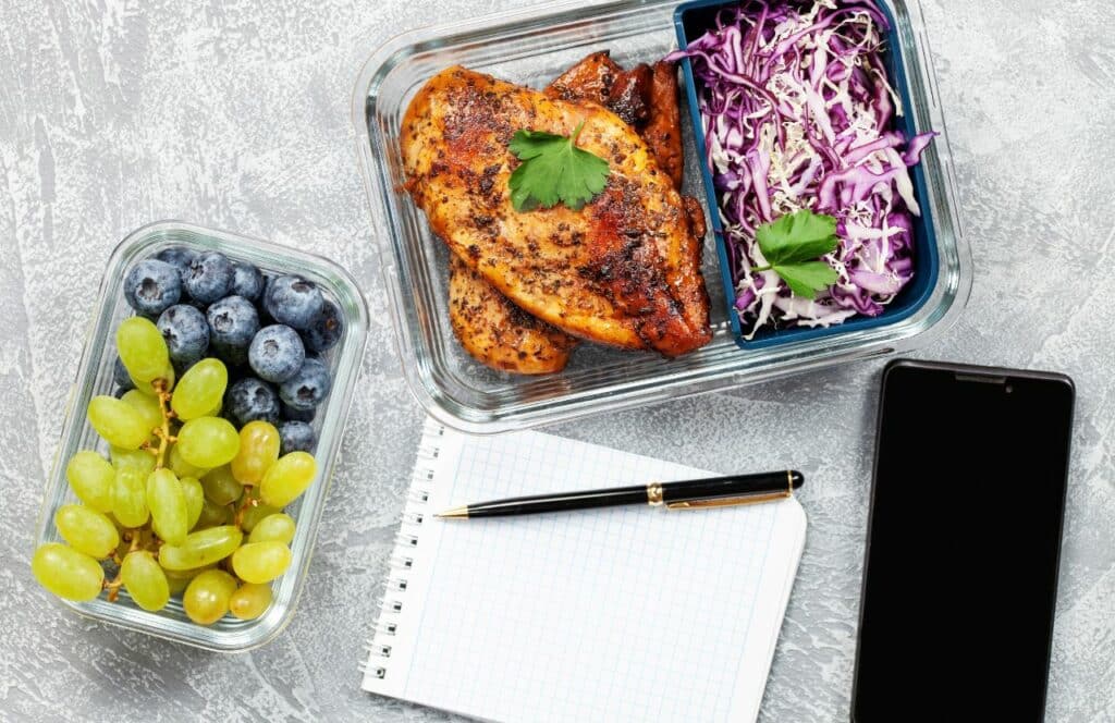 A chicken and cabbage meal, with fruit packaged up and ready to eat on a table with a notepad, pen, and phone. Keep reading for tips on how to write blog posts faster.
