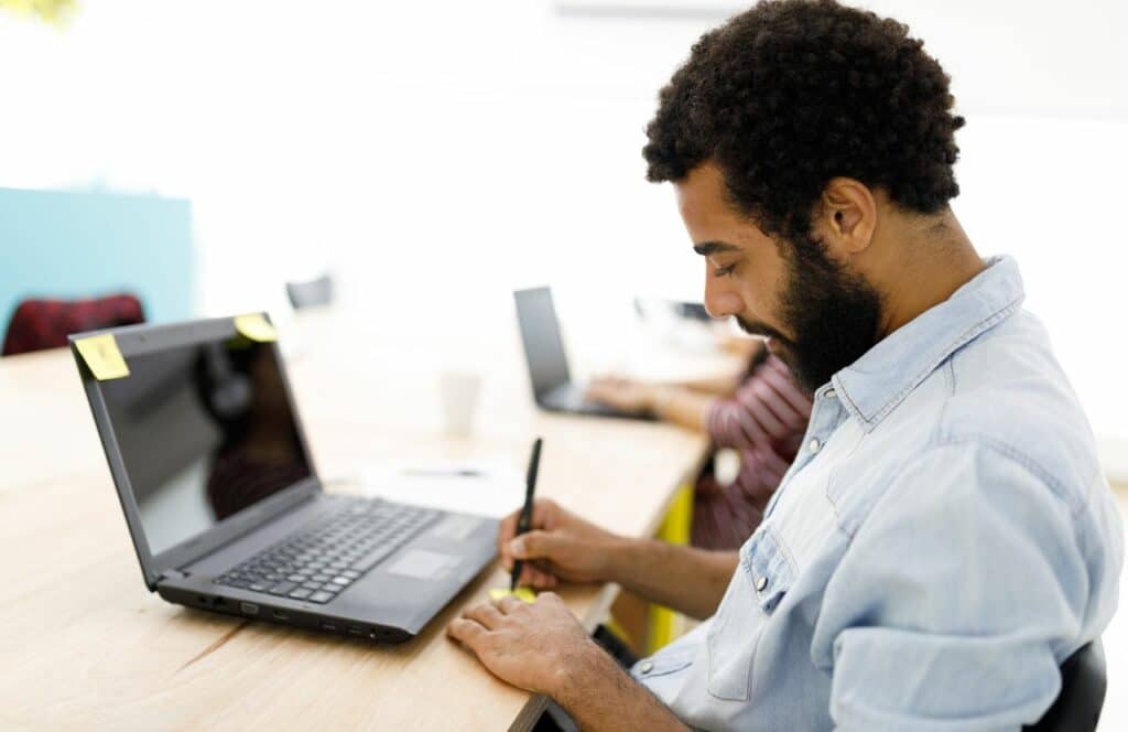 Black man sitting at a desk taking notes to prepare for writing blog posts. Keep reading for tips on how to write blog posts faster.