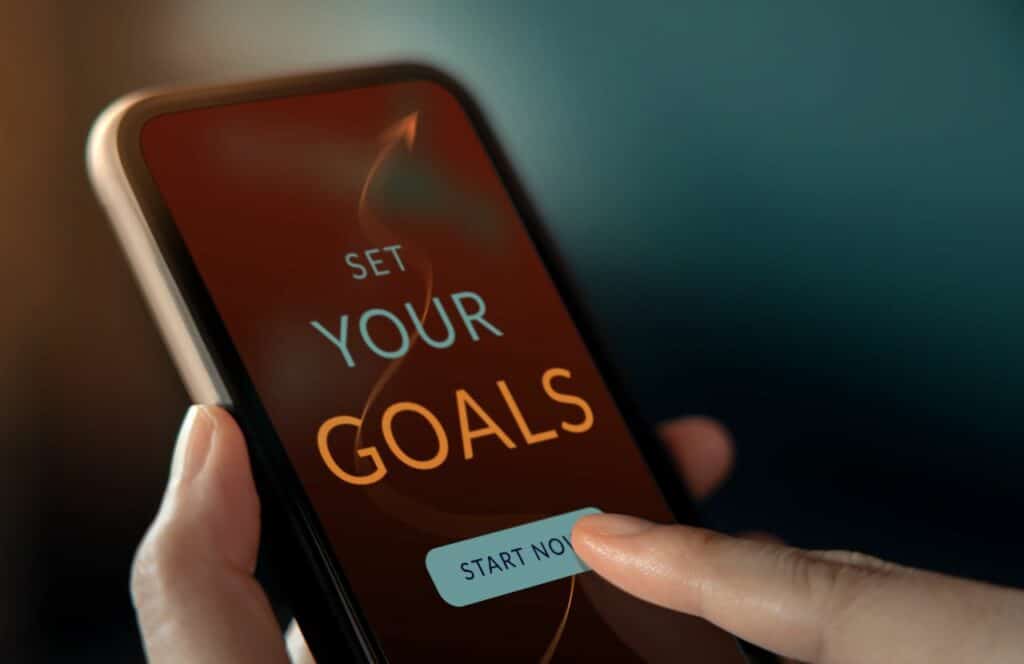 Person holding a phone with a personal goal setting app. Keep reading for tips on how to write blog posts faster.