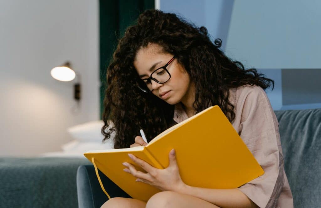 Young woman with curly hair and glasses writing in a yellow journal to document her progress with setting personal goals. Keep reading for tips on how to write blog posts faster.