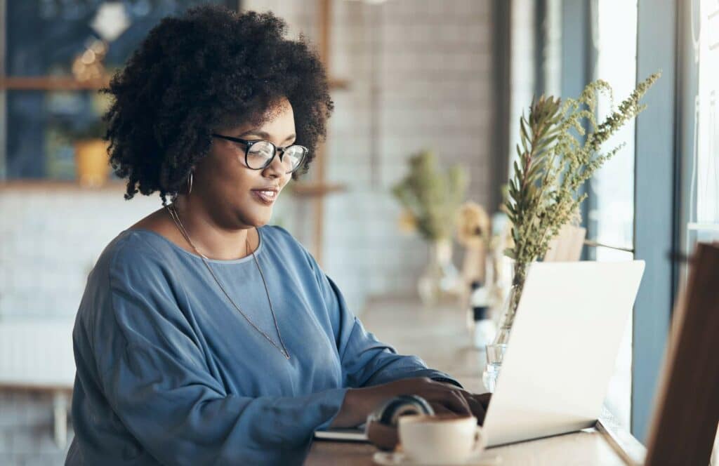 A black woman with a blue top, sitting with her laptop creating a digital product for her blog. Keep reading to learn how to make money blogging.