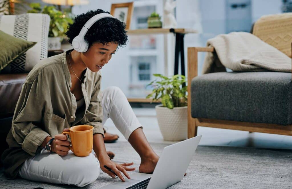 A woman with white earphones, sitting on the floor with a cup of coffee while writing on her blog. Keep reading to learn how to monetize your blog from day one.