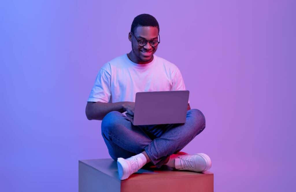 A black man sitting on top of a box in a purple room with his laptop as he creates blog posts. If you have ever asked "What's the point of blogging?" click here to read more.