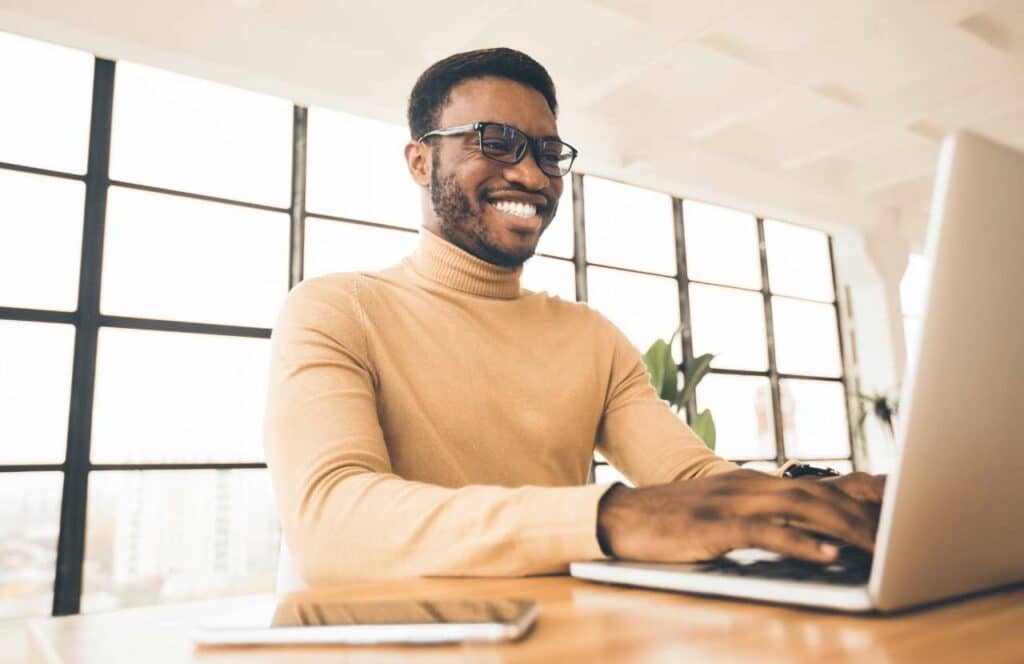 A black man with a light brown turtle neck on and smiling while blogging in an office space because his blog traffic is increasing. If you have ever asked "What's the point of blogging?" click here to read more.
