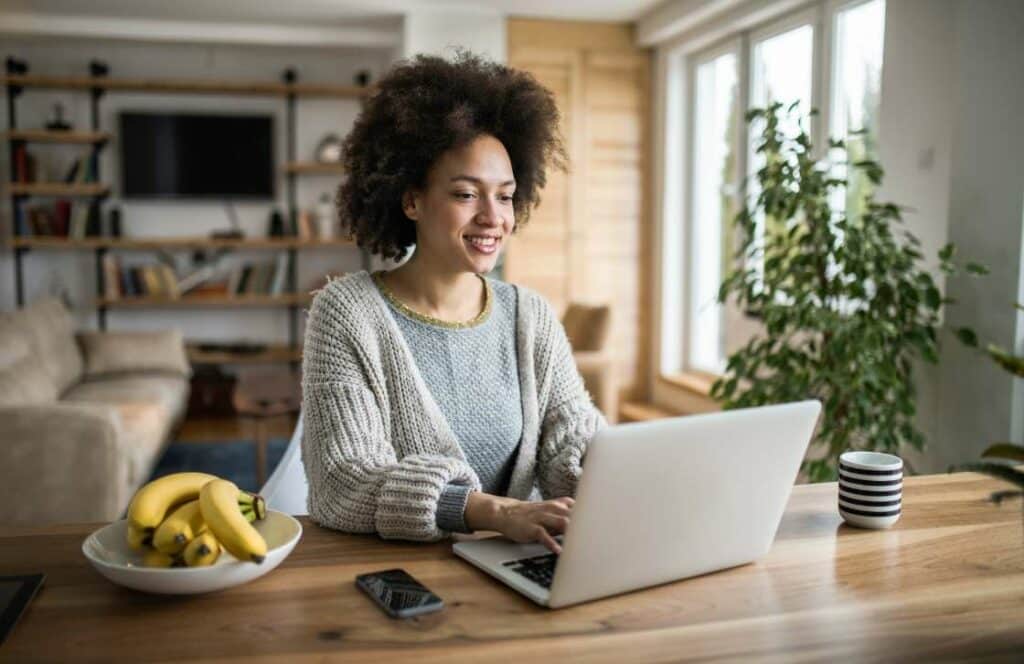 A black woman sitting at a table next to a bowl of bananas and a coffee cup while publishing blog posts. If you ever wondered "What's the point of blogging?" click here to read more.