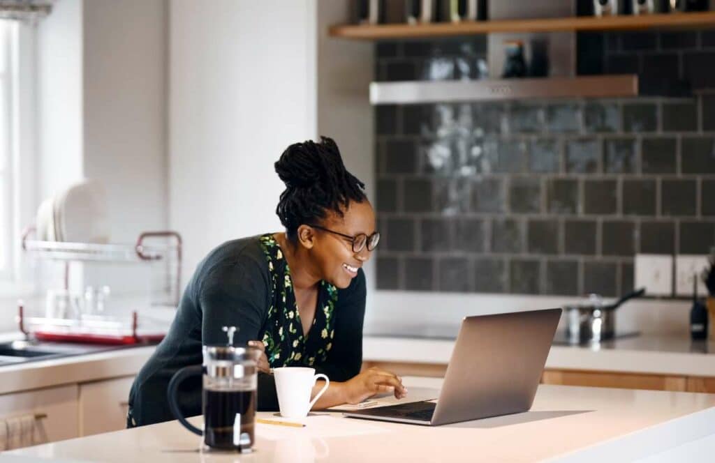 A black woman standing at the kitchen bar with coffee and a laptop as she works on her blog. If you have ever asked "What's the point of blogging?" click here to read more.