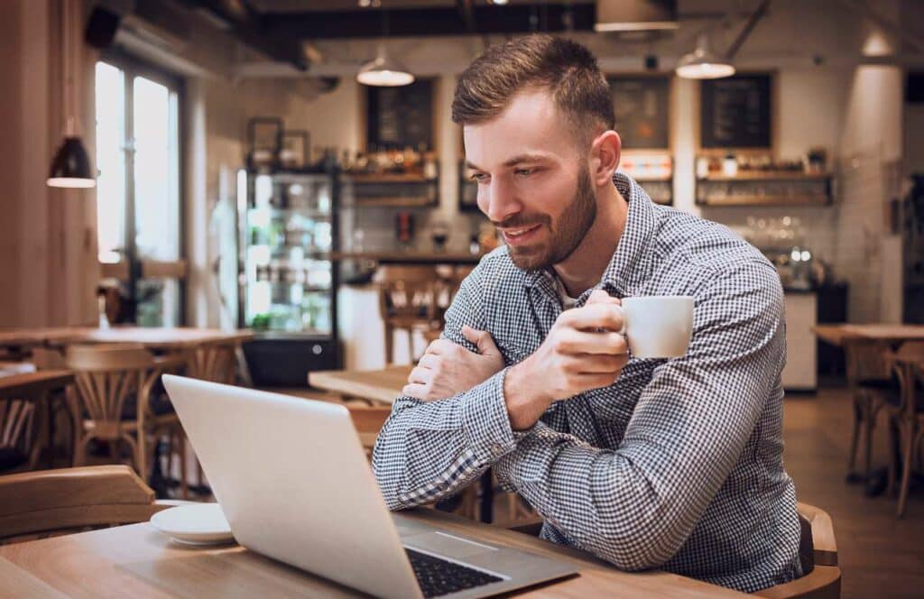 A man sitting in a coffee shop with a cup of coffee and his laptop while working on his next blog post. If you have ever asked "What's the point of blogging?" click here to read more.
