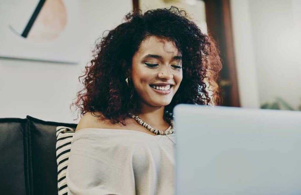 A woman sitting and smiling on the couch while creating her weekly blog post which are increasing her website traffic. If you have ever asked "What's the point of blogging?" click here to read more.