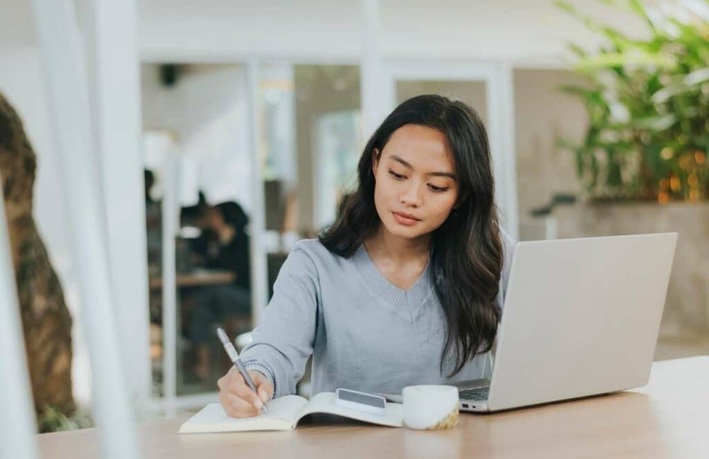 A woman sitting at a desk with a phone, laptop, journal, and pen so she can create blogging goals. Keep reading to learn more about how you can increase website conversions.