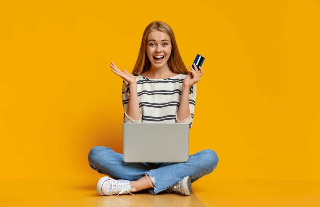 A woman smiling and sitting on the floor with her laptop while holding a credit card as she makes a one-time purchase on a blogger's website. Learn more about conversion rate optimization for your blog.