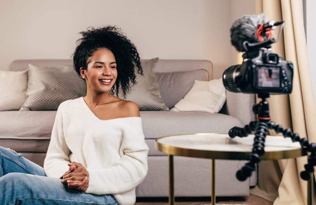 A woman with long curly hair sitting in front of the couch with a camera while recording content for her blog. If you have ever asked "What's the point of blogging?" click here to read more.