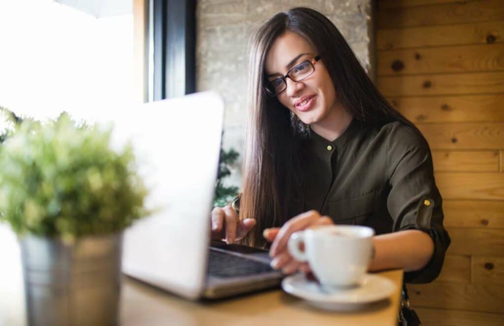 A woman with long hair sitting with a cup of coffee and her laptop while working on her next blog post. If you have ever asked "What's the point of blogging?" click here to read more.