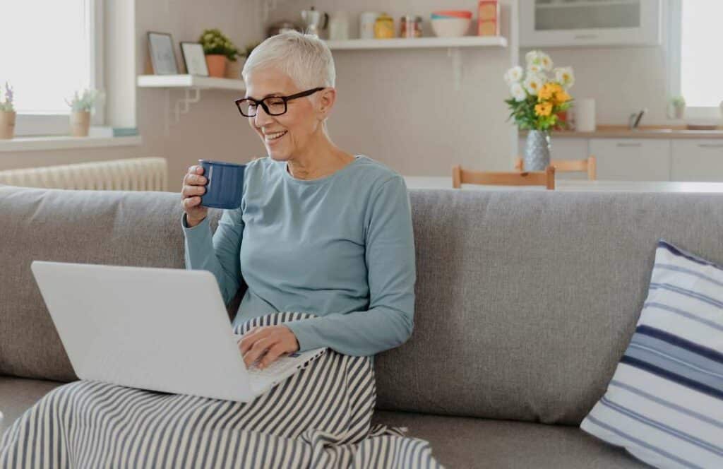 An older woman with short hair holding a blue coffee mug while sitting on the couch and creating a blog post. If you have ever asked "What's the point of blogging?" click here to read more.