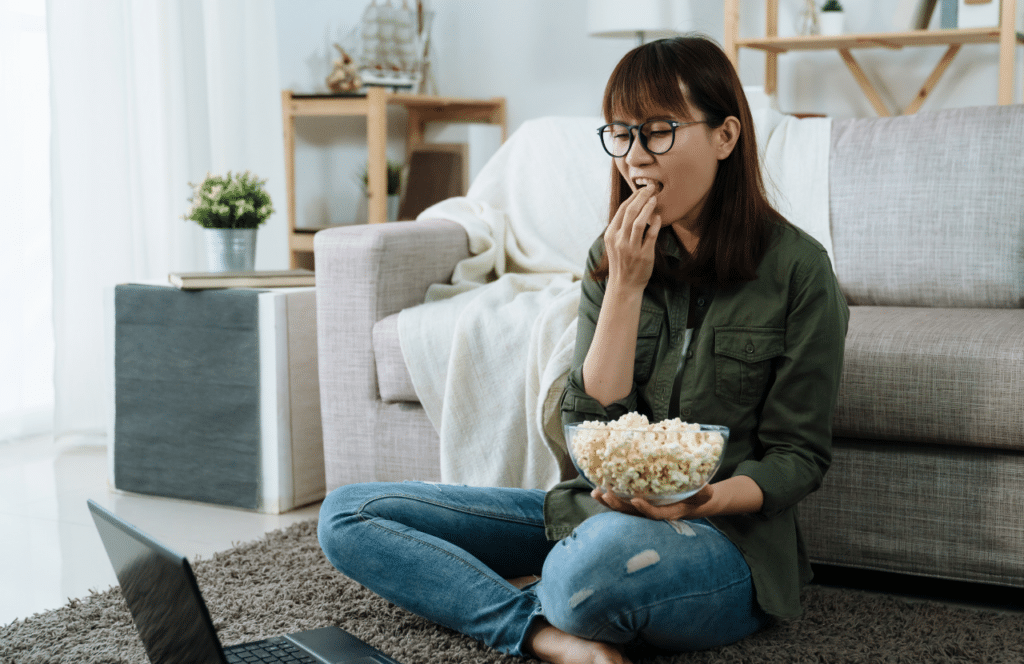 Woman sitting on the floor in front of couch with laptop and popcorn as she gets ready to write her weekly blog post. Keep reading to learn the answer to the question, "Is Blogging Still Relevant?"