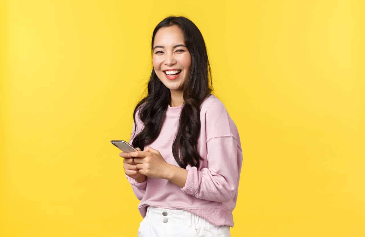 A smiling woman standing in front of a yellow background with her phone as she reads tips about blog brand awareness