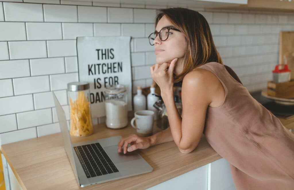A woman on her laptop in the kitchen thinking about if she should use an ai blog writer for her blog.